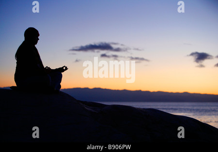 Eine junge Frau mit einem Hut macht Yoga und meditiert im Freien auf einem Felsen am Meer. Stockfoto