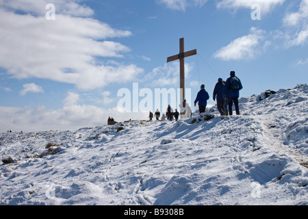 Menschen (Wanderer), die auf schneebedeckten Hügeln zu hohen hölzernen Osterkreuz (Symbol des christlichen Glaubens) - Otley Chevin, West Yorkshire, England, Großbritannien. Stockfoto