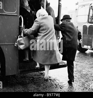 1950s, im Winter, knabern ein älterer Mann und eine ältere Frau die Handgeländer und steigen auf die offene hintere Plattform eines Doppeldeckerbusses, London, England, Großbritannien. Stockfoto