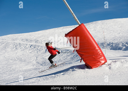 Eine junge Frau in einer roten Skijacke springt über einen Anstieg der Piste hinter einer roten Sperr-Matratze. Schneebedeckte Hügel und blauer Himmel Stockfoto