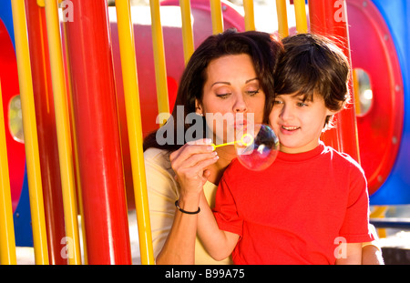 Hispanische Mutter und Sohn Spaß Seifenblasen und umarmt auf bunte Ausstattung im Park im freien Stockfoto