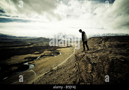 Ein Mann schaut die Felswand ein Lava Butte mit Blick auf einen grasbewachsenen Tal mit schneebedeckten Gipfeln im Hintergrund. Stockfoto