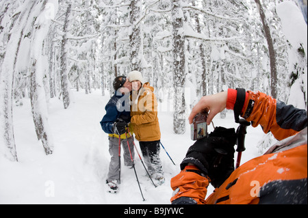 Eine Mitte Erwachsene Frau und Mann Pose während eine junge Frau ihr Bild beim Schneeschuhwandern durch ein Schnee nimmt bedeckt Wald. Stockfoto