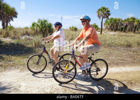 Älteres Paar Reiten am Strand mit Sonnenbrille Fahrräder und Helme Fokus auf die Frau Stockfoto