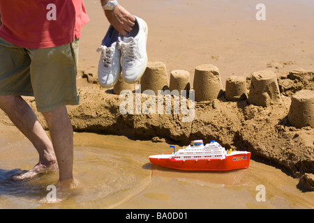 Ein Mann in kurzen Hosen mit seinen Schuhen in der Hand geht, Weg von einem Spielzeug-Kreuzfahrtschiff schwebend in den Graben eine große Sandburg Stockfoto
