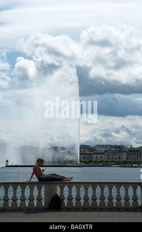 Eine Mädchen sitzt auf einer Mauer mit Blick auf den Springbrunnen Jet d ' Eau in Genf, während ihr Mobiltelefon tragen ein rotes top Stockfoto