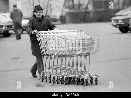 Achtziger Jahre, schwarz / weiß Foto, Wirtschaft, Einzelhandel, Einkaufszentrum, älterer Mann schiebt mehrere shopping Wagen über einen Parkplatz an der Sammelstelle im Alter von 65 bis 70 Jahre Stockfoto