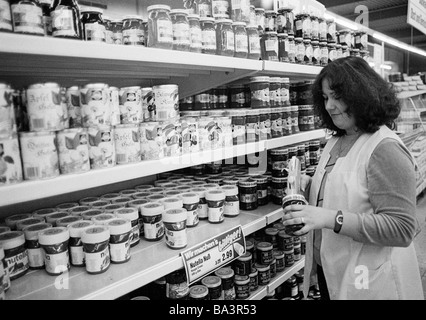 Achtziger, schwarz / weiß Foto Beschäftigten Leben, Einzelhandel, Shop, Köstlichkeiten, Verkäuferin, junge Frau in einem Mantel Arbeit steht in der Nähe ein Supermarkt-Regal und Preise die waren durch eine Etikettiermaschine, im Alter von 23 bis 30 Jahre Stockfoto