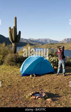 Ein Mann gähnt und erstreckt sich beim camping allein bei Sonnenaufgang auf einer Klippe mit Blick auf Apache Lake Arizona Stockfoto