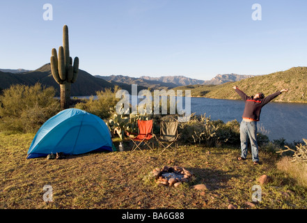 Ein Mann gähnt und erstreckt sich beim camping allein bei Sonnenaufgang auf einer Klippe mit Blick auf Apache Lake Arizona Stockfoto