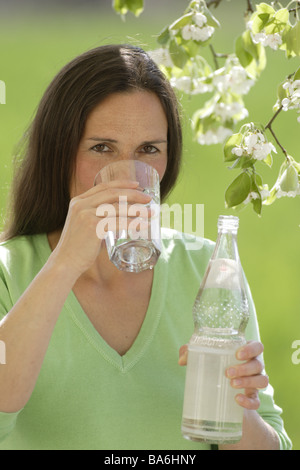 Frau langhaarige Brünette Mineralwasser Getränke Blick Kamera Frühling draußen Porträt 30-40 Jahre Garten Baum blüht Obstbaum Stockfoto