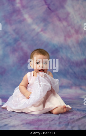 Ein sechs Monate altes Baby Mädchen im rosa Kleid sitzt vor einem rosa, lila und blau Hintergrund halten ihr Kleid bis zu Ihrem Stockfoto