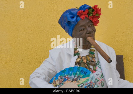 Ältere Frau, die eine große Zigarre rauchend in die weiß gekleidet der Santeria mit einem Kopfschmuck und Blumen sowie ein Fan. Stockfoto