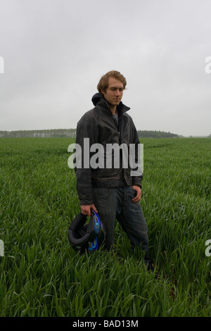 Ein junger Mann trägt eine schwarze Lederjacke, hält einen Motorradhelm Stand in einem grünen Feld Stockfoto