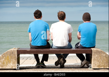 Rückansicht der drei jungen Männer tragen T-shirts auf einer Bank sitzen und Blick aufs Meer Aberystwyth Promenade Wales UK Stockfoto