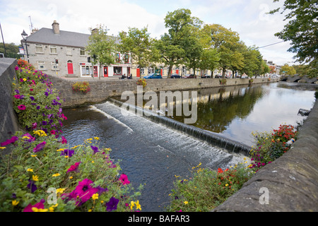 Wehr am Fluss Carrowbeg. Ein kleiner Wasserfall entsteht durch das Wehr auf dem Blumen gesäumten Fluss im Zentrum von Westport. Stockfoto