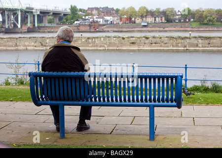 Ein einsamer Mann sitzt auf einem blauen Sitzplatz mit Blick auf das Silberjubiläum-Brücke über den Fluss Mersey und Manchester Ship Canal Stockfoto