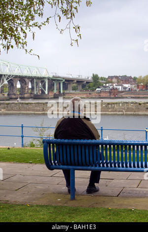 Ein einsamer Mann sitzt auf einem blauen Sitzplatz mit Blick auf das Silberjubiläum-Brücke über den Fluss Mersey und Manchester Ship Canal Stockfoto