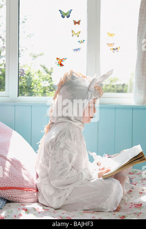 Junge Mädchen (5-6) sitzen im Bett im Einhorn Kostüm Lesebuch, Seitenansicht Stockfoto