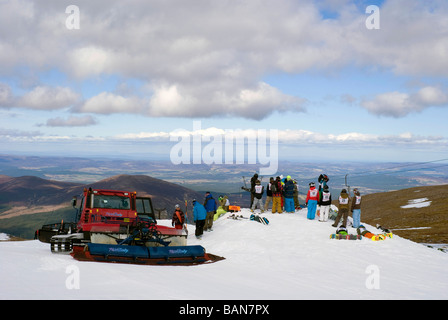 Piste-Beißer Maschine arbeitet auf Cairngorm Berg, in der Nähe eine Gruppe von jungen Snowboarder wartet auf Rennen Stockfoto