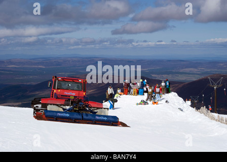 Piste-Beißer Maschine arbeitet auf Cairngorm Berg, in der Nähe eine Gruppe von jungen Snowboarder wartet auf Rennen Stockfoto