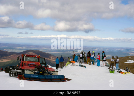 Piste-Beißer Maschine arbeitet auf Cairngorm Berg, in der Nähe eine Gruppe von jungen Snowboarder wartet auf Rennen Stockfoto