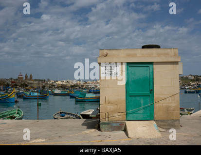 Blauer Himmel und den malerischen Hafen von maltesischen Luzzu (Fischerboote) hinter einem kleinen Gebäude in Marsaxlokk, Malta Stockfoto