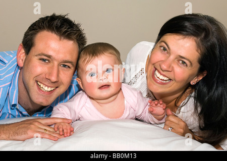 Horizontale Nahaufnahme Portrait von einem niedlichen sechs Monate alten Babymädchen und ihren stolzen Eltern liegend auf einem Bett, Blick nach oben Stockfoto