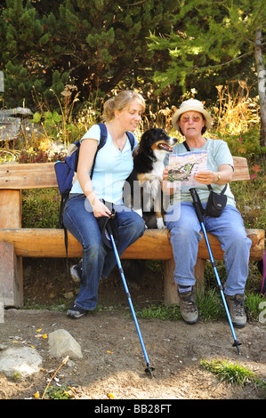 Zwei Frauen Wanderer sitzt auf einer Holzbank, eine Pause, mit ihrem Hund zwischen ihnen. Stockfoto