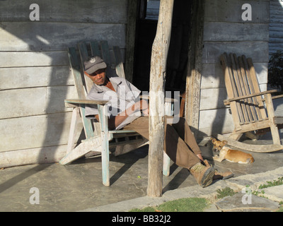 älteren kubanischen arbeitenden Menschen Entspannung in Holzstuhl auf sonnigen Deck einfaches Holzhaus mit kleiner Hund und leere Schaukelstuhl Stockfoto