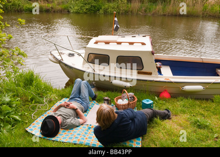 Ein Ehepaar mittleren Alters, ein Picknick am Flussufer neben ihrem Boot auf dem Fluß Waveney auf den Norfolk Broads im Vereinigten Königreich Stockfoto