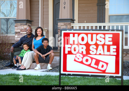 Afroamerikanische Familie sitzen zusammen auf der Eingangstreppe des Hauses mit Immobilien Schild im Vordergrund Stockfoto