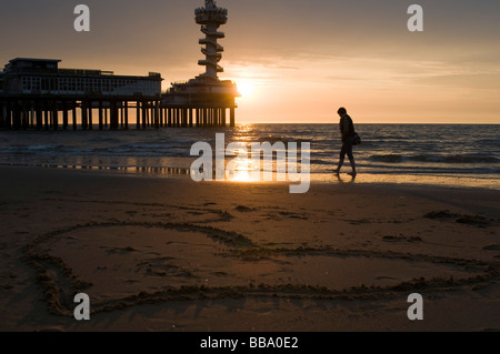 Frau allein zu Fuß am Strand bei Sonnenuntergang mit Loveheart gezeichnet in Sand und Pier im Hintergrund, Scheveningen, Niederlande Stockfoto