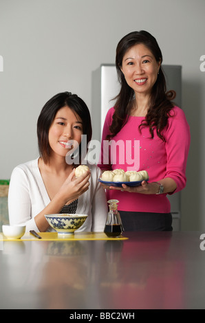 Mutter und Tochter in der Küche, jüngere Frau sitzen, ältere Frau stand neben ihr hielt einen Teller mit chinesischen Brötchen Stockfoto