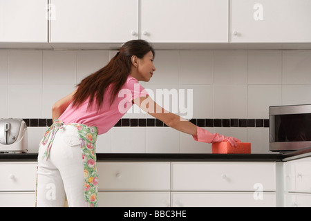 Frau stehend in Küche, tragen von Handschuhen und Schürze und Zähler mit großen rosa Schwamm reinigen. Stockfoto
