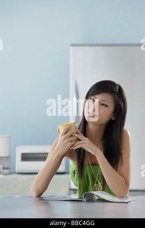 Frau sitzt in der Küche mit Magazin Holding Becher Stockfoto