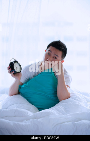 Mann sitzt im Bett, mit Wecker, Hand am Kinn Stockfoto