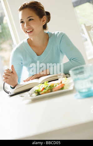 Junge Frau sitzt am Tisch, Salat auf Tisch, schreiben in Notebook, wegschauen Stockfoto