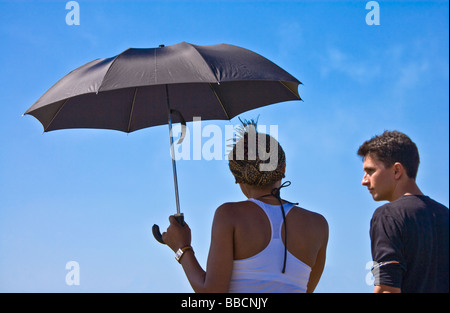 Rückansicht des jungen schwarzen Frau Beschattung selbst von der Sonne unter einem schwarzen Schirm mit ein junger weißer Mann sah Sie Stockfoto