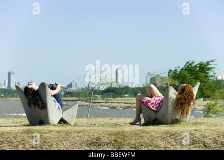 Zwei junge Frauen, die zum Entspannen in Stühle, Teich und Stadt Skyline im Hintergrund Stockfoto