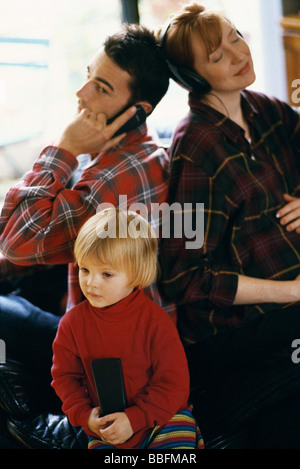 Familie sitzen Rücken an Rücken mit Handy, Mutter Tochter vor dem Fernseher, Kopfhörer, hören, Vater Stockfoto