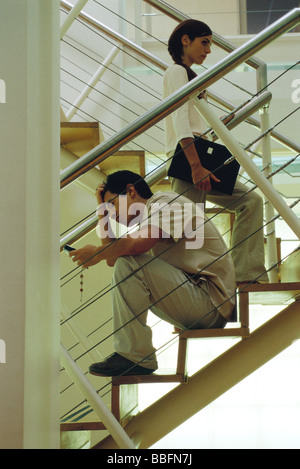 Mann mit Hand am Kopf sitzt auf der Treppe, Blick auf Handy, Frau vorbeigehen Stockfoto