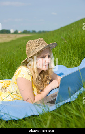 Blondes Mädchen mit Hut, lächelnd und liegend auf einer Decke auf einer Wiese an einem Notebook arbeitet Stockfoto