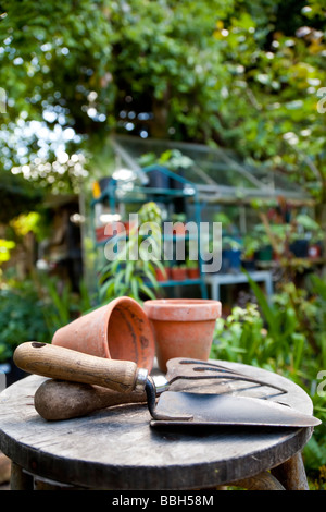 Gartenarbeit, Utensilien und Blumentöpfe ruht auf einem Hocker in einem grünen Garten mit einem Gewächshaus im Hintergrund unscharf Stockfoto