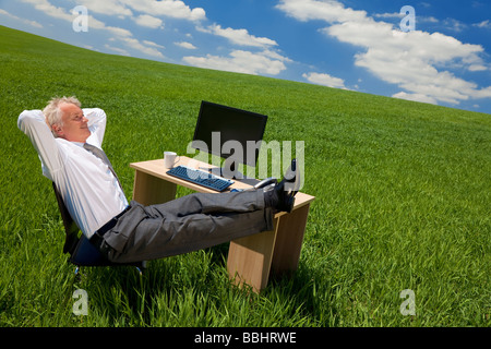 Business Konzept Schuss eines Geschäftsmannes entspannend mit seinen Füßen oben auf seinem Schreibtisch in einem grünen Feld mit einem strahlend blauen Himmel und Wolken Stockfoto