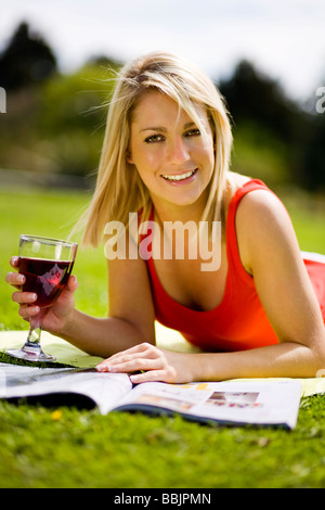 Eine junge Frau sonnt sich im Park beim Lesen einer Zeitschrift und ein Glas Wein zu genießen. Stockfoto