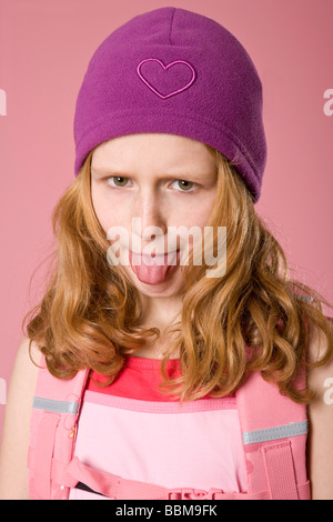 Rothaarige Mädchen tragen eine violette Motorhaube und eine Schultasche vor einem rosa Hintergrund, stossen ihre Zunge Stockfoto