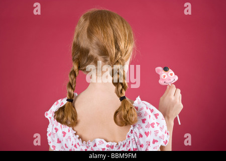 Rothaariges Mädchen mit Zöpfen tragen ein Sommerkleid vor einem roten Hintergrund mit einem Lutscher Stockfoto