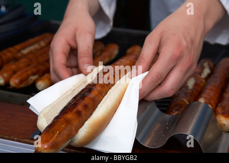 Kreditor Vorbereitung Bratwurst in Brot Roll Bap auf Serviette mit deutschen Würstchen auf dem Grill zum Verkauf an ein outdoor-Markt in Großbritannien Stockfoto