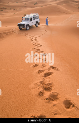 Ein Mann in traditioneller Kleidung der Berber geht zu seinem Auto in der Nähe von Merzouga Marokko Nordafrika durch die Dünen der Wüste Erg Chebi Stockfoto